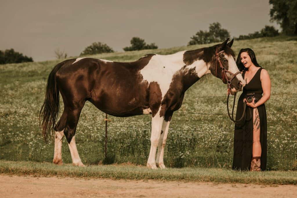 A woman in a long black dress holding a harness on a brown and white horse. She is looking at the horse and smiling.