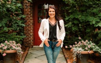 A woman in jeans and a white blazer standing in front of a house.