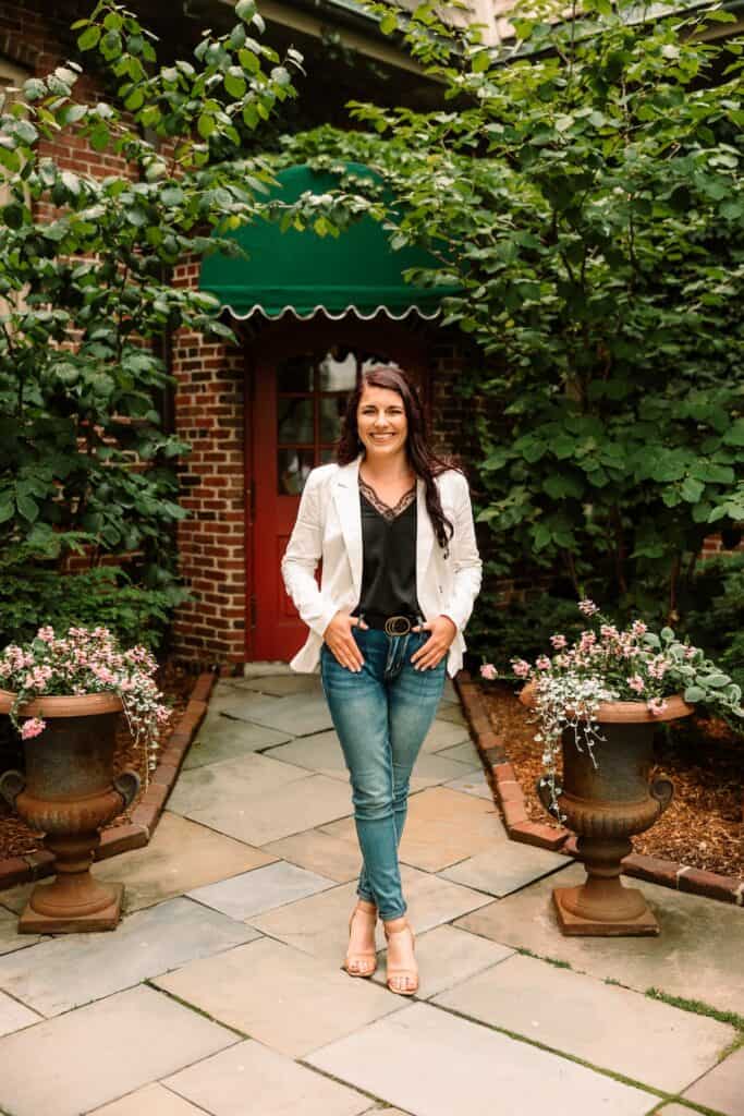 A woman in jeans and a white blazer standing in front of a house.