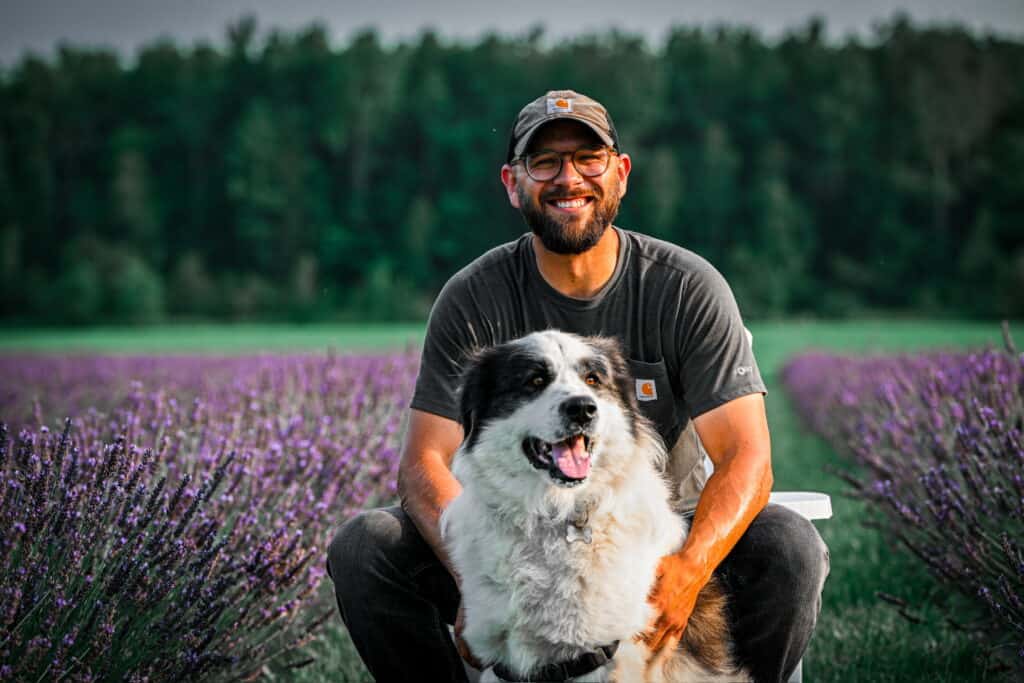A man with a beard and hat sitting on a bench with his dog in a lavender field.